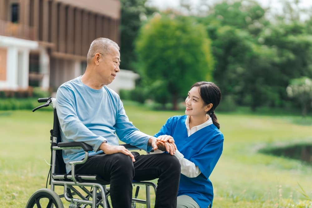 A nurse holds an elderly man’s hand while kneeling beside his wheelchair while enjoying the outdoors.